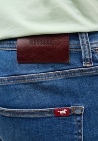 Niebieskie dżinsy z pomarańczowymi szwami, z bordową skórzaną metką z napisem "MUSTANG" oraz małą czerwoną metką z białym emblematem konia.