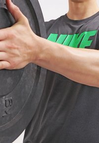 Czarna talerz do ciężarów trzymany w ręku przez osobę w jasnej, teksturowanej, szarej koszulce z jaskrawozielonym logo. Zbliżenie na muskularną rękę i oznaczenia talerza.