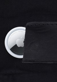 Apple AirTag delvist sat ind i en sort læderlomme på en mørk stofbaggrund, der viser Apple-logoet og tekst rundt om kanten.