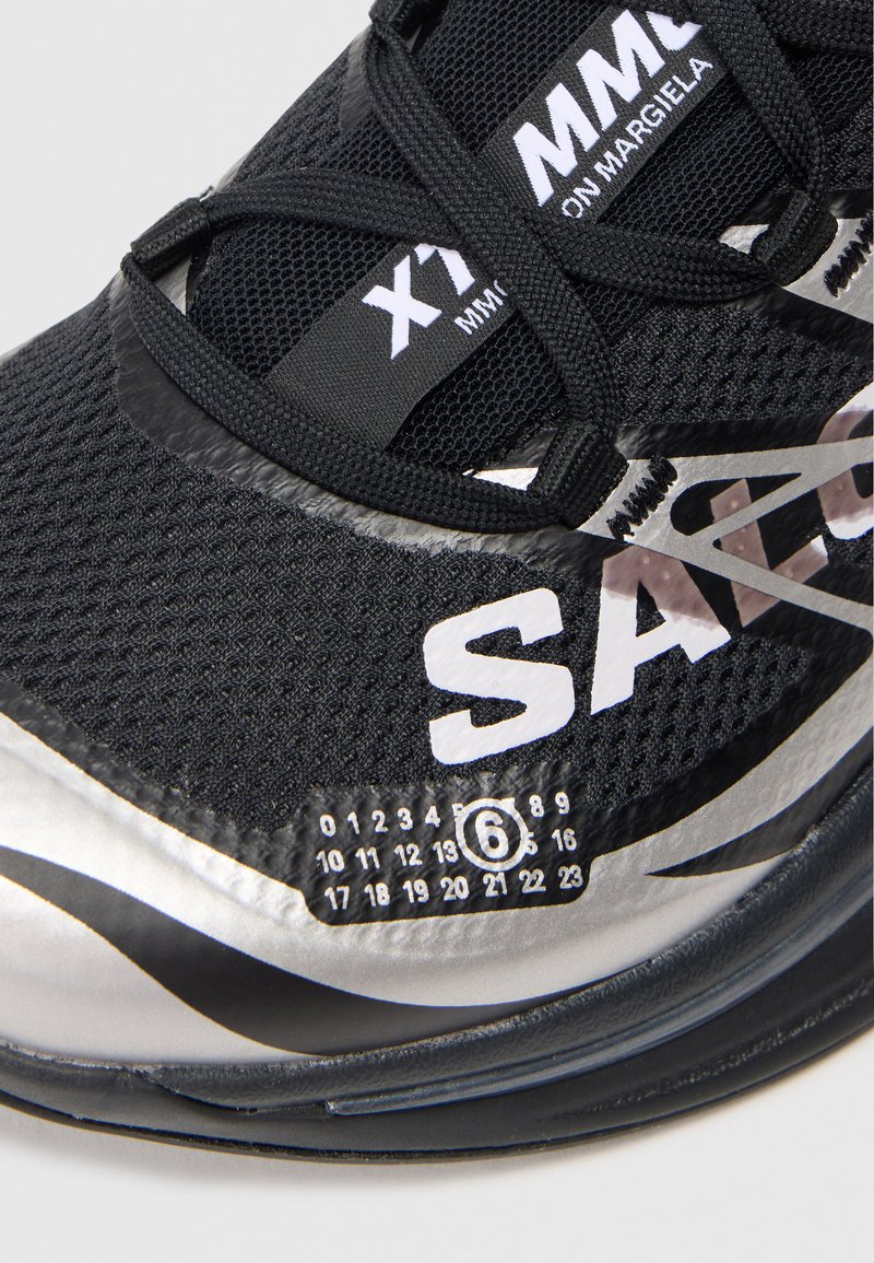 Sort sneaker lavet af mesh og syntetiske materialer, med et strømlinet design, hvid branding og en unik nummereret skala-detalje på siden.