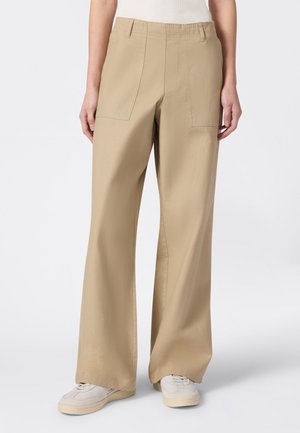 Beżowe spodnie o szerokich nogawkach z przednimi kieszeniami naszywanymi, noszone z białymi trampkami i włożoną do środka jasną górą, stojące na jednolitym tle.