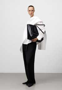 Donna con camicia bianca oversize e pantaloni neri larghi che tiene una grande borsa tote in pelle nera, in piedi davanti a uno sfondo chiaro uniforme.