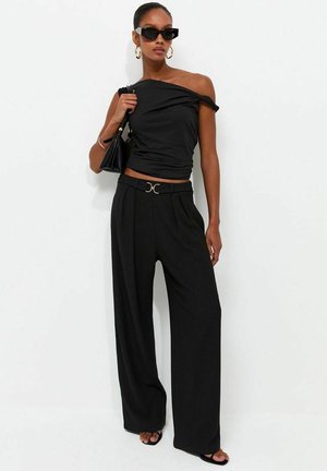 Kvinna som bär svart off-shoulder topp, vida byxor med hög midja, svarta sandaler, solglasögon och guldfärgade hoops örhängen, håller en svart handväska.