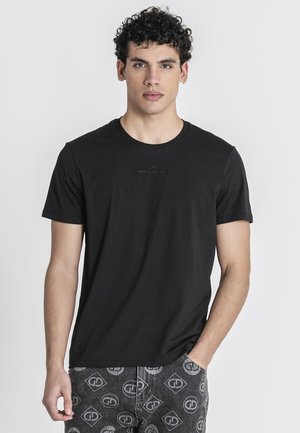 Joven con cabello rizado que lleva una camiseta negra lisa de manga corta y pantalones gris oscuro estampados sobre un fondo claro.