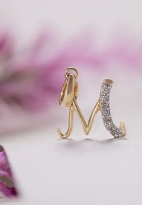 Charm en forme de lettre "M" en or, avec une finition lisse et un design incurvé, accentué par des strass transparents d'un côté.