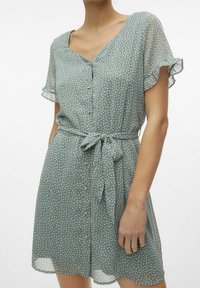 Grünes Polka-Dot-Kleid mit V-Ausschnitt, kurzen, flatternden Ärmeln, vorderen Knöpfen und einem Knoten-Gürtel in der Taille. Leichtes, transparentes Material.