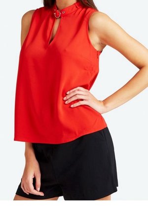 Ærmeløs rød top med nøglehuludskæring og ringdetalje, kombineret med højtaljede sorte shorts på en person med den ene hånd på hoften.