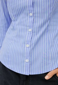 Jasnoniebieska koszula z guzikami w paski, z białymi pionowymi paskami, charakteryzująca się gładką tkaniną, zapięciem na guziki i zaokrąglonym dołem.