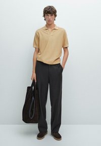 Polo shirt beige a maniche corte con finitura testurizzata, abbinato a pantaloni scuri su misura e una borsa tote in suede marrone. Calzature casual completano il look.