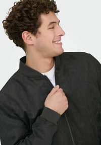 Jeune homme souriant, tenant la fermeture éclair d'une veste bomber noire par-dessus une chemise blanche, regardant vers la droite sur un fond clair.