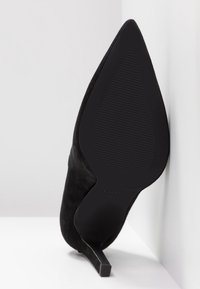 Zapato de tacón alto de ante negro con punta afilada y un tacón de aguja delgado. Presenta una suela de goma negra texturizada y un diseño minimalista.