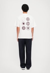 Hvit T-skjorte med kunstneriske grafikker i mørk maroon på baksiden, sammen med løstsittende bukser med mørke striper og hvite joggesko.