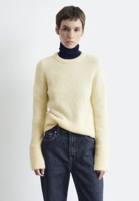 Gelbes flauschiges Sweatshirt mit langen gerippten Bündchen, über einem marineblauen Rollkragenpullover getragen. Kombiniert mit dunkelblauen Jeans, die traditionelle Taschen haben.