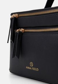 Bolso de mano sintético negro con dos cremalleras doradas, con una textura suave y un logo en relieve que dice "ANNA FIELD" en la parte delantera.