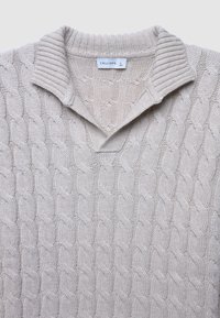 Beige gestrickter Pullover mit einem Kabelmuster, einem spitzen Kragen und strukturierten Rippakzenten an den Bündchen und dem Kragen. Größenschild sichtbar.