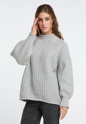 Junge Frau mit langen Haaren, die einen lockeren grauen Strickpullover und dunkle Jeans trägt, steht vor einem einfarbigen hellen Hintergrund.