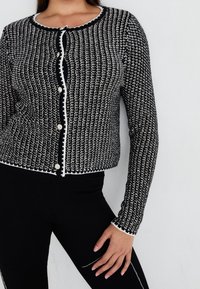 Cardigan tricoté noir et blanc avec un col rond, une fermeture à boutons et une bordure contrastante. Présente un motif texturé et des manches longues.