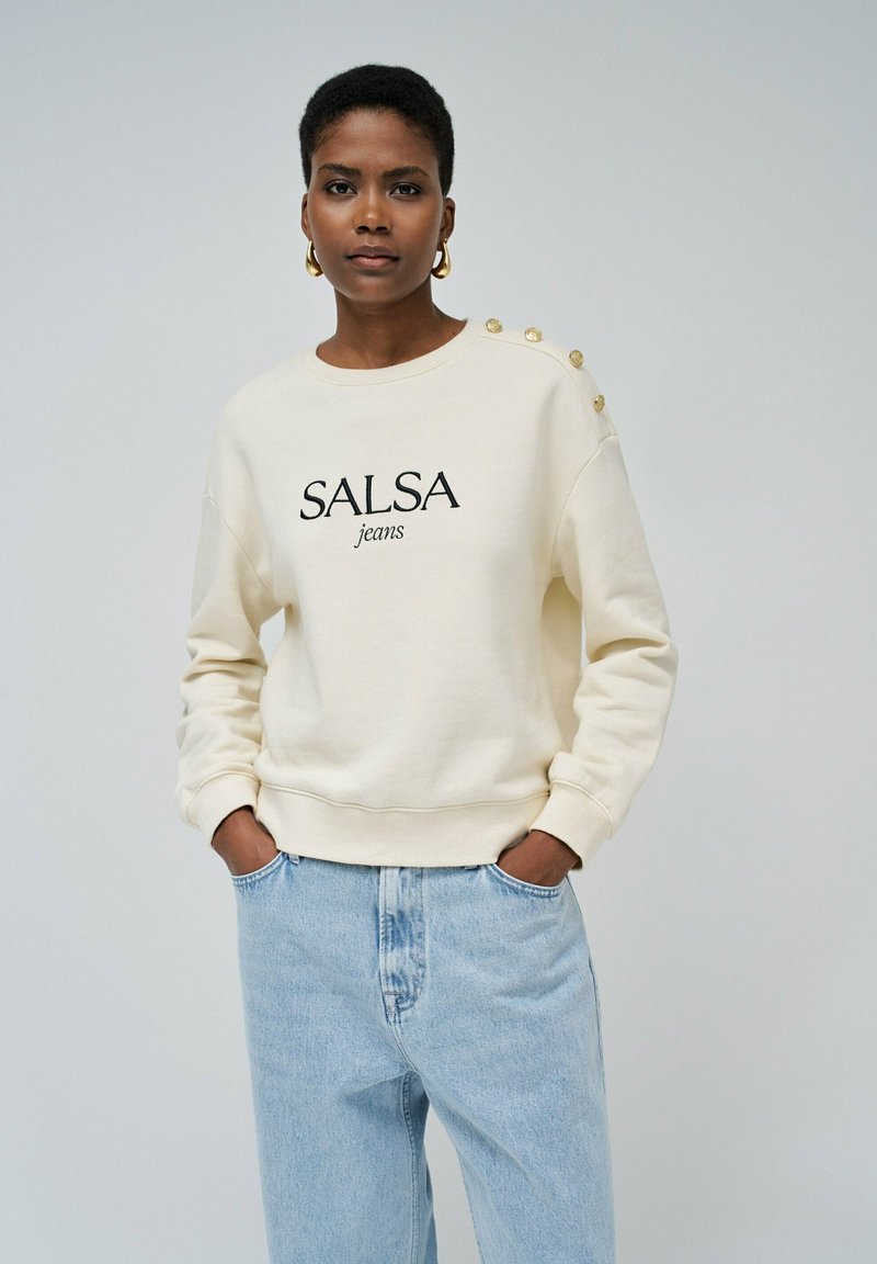 Světlý krémový svetr s potiskem "SALSA jeans" v černé barvě, se zlatými knoflíky na levém rameni a žebrovanými manžetami.