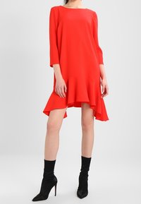 Vestido rojo de manga larga con un dobladillo asimétrico y volantes acampanados. Textura suave, diseño minimalista, combinado con botas de tacón de aguja negras.