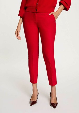 Femme portant un pantalon rouge vif ajusté longueur cheville et un cardigan rouge assorti avec des boutons en forme de cœur, ainsi que des escarpins bordeaux à bout pointu.