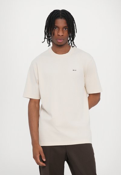 Camiseta color crema de algodón suave, con un corte clásico de cuello redondo y mangas cortas. Texto "NN. 07" impreso en el lado izquierdo del pecho.