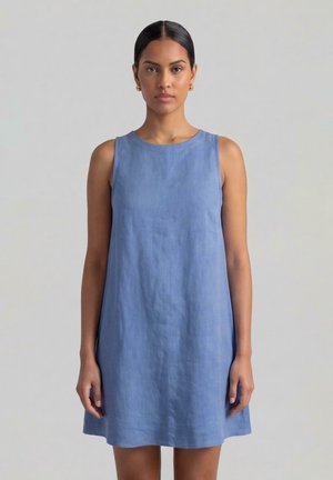 Donna con capelli scuri raccolti indietro, che indossa orecchini a cerchio dorati e un vestito di lino blu senza maniche e ampio, in piedi davanti a uno sfondo chiaro e semplice.
