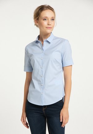 Giovane donna che indossa una camicia azzurro chiaro a maniche corte con bottoni e jeans scuri, in piedi davanti a uno sfondo bianco semplice.