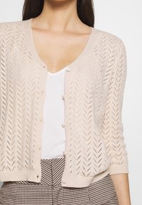 Ljust beige stickad cardigan med ett zick-zack mönster, korta ärmar och knäppning. Bärs över en vit topp och rutiga byxor.