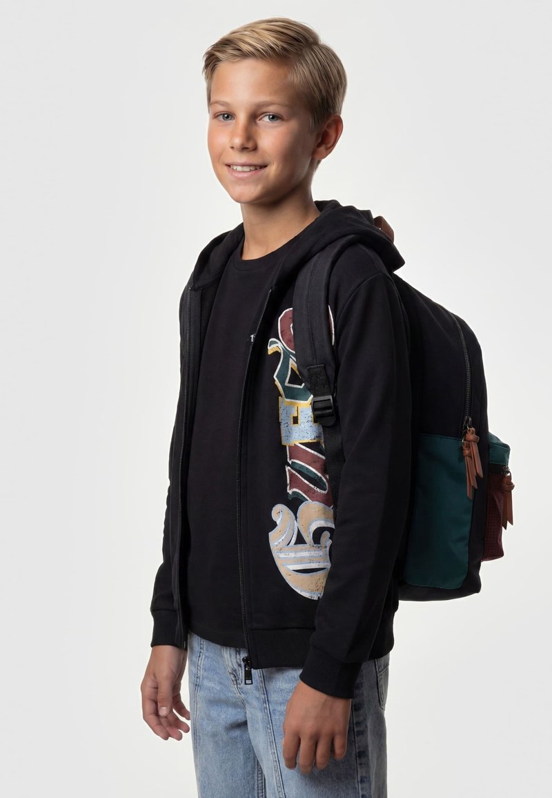 Garçon souriant aux cheveux blonds courts portant un sweat à capuche noir, un t-shirt graphique, un jean clair et un sac à dos noir et vert.
