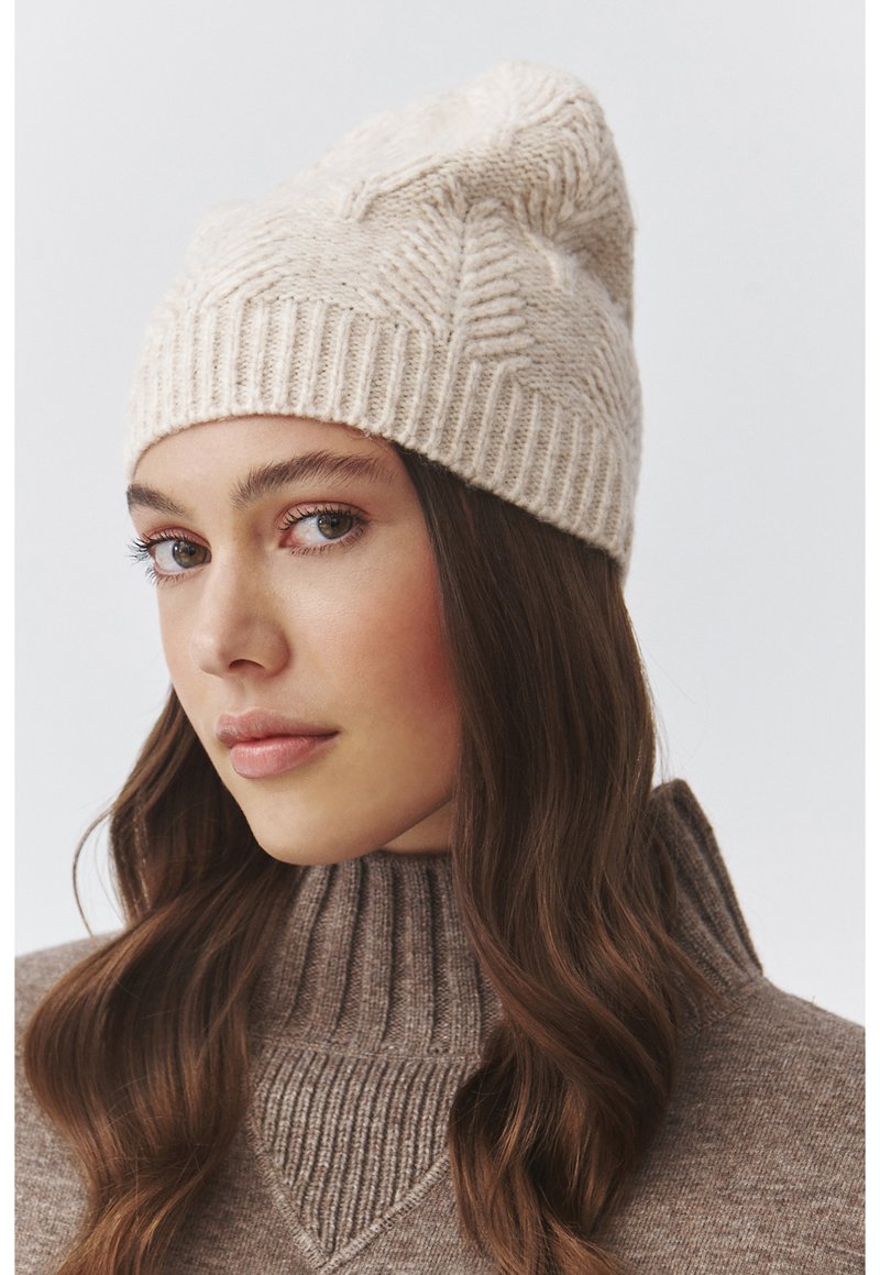 Gorro beige de punto con un patrón texturizado y un ajuste holgado, combinado con un suéter marrón de cuello alto y con canalé.