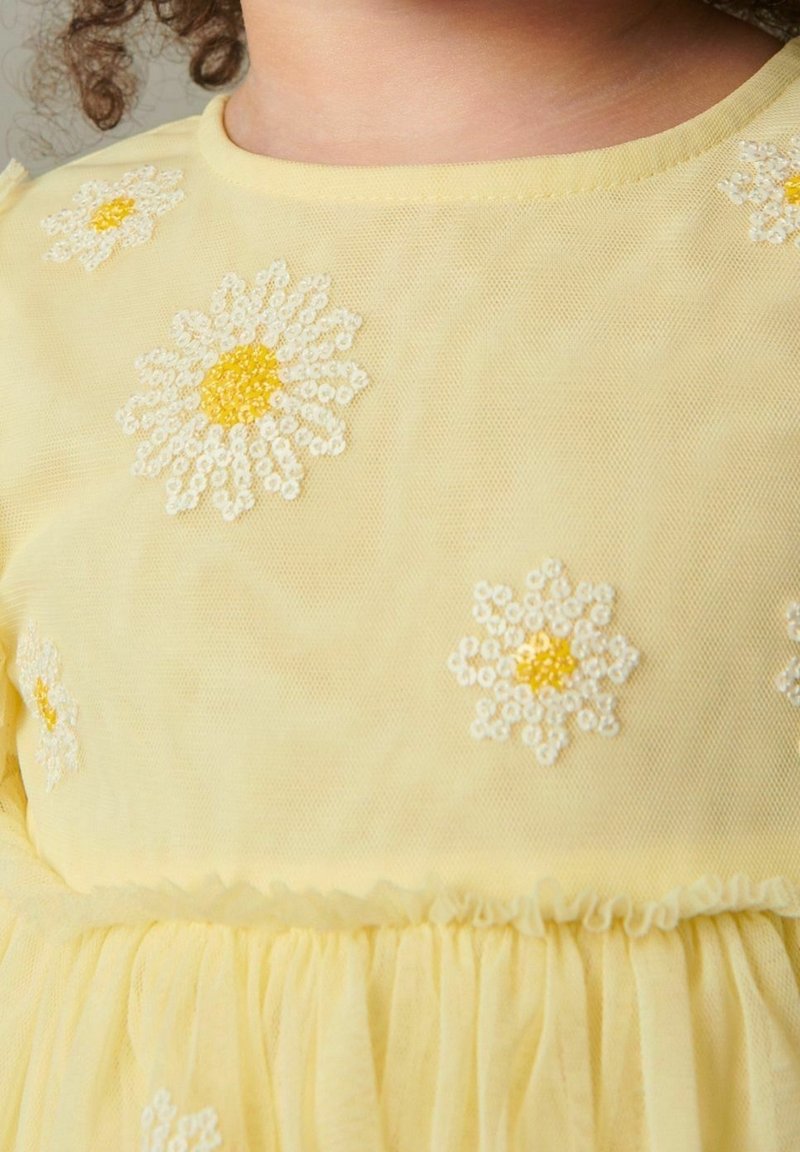 Robe jaune ornée de broderies florales avec des pétales blancs et des centres jaunes, en tissu transparent avec un détail de jupe froncée.