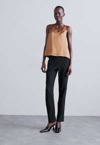 Canotta in satin marrone abbinata a pantaloni plissettati neri e tacchi neri lucidi, caratterizzata da un design moderno e slanciato con una silhouette aderente.