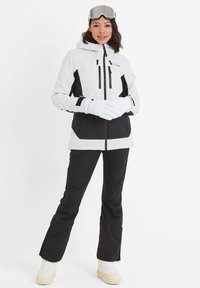 Ski jacket in white and black, featuring a hood, multiple zip pockets, and a sporty design. Paired with black ski pants and gloves.