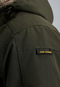 Donkergroene jasdetail met een gestructureerde stof, zichtbare stiksels en een zwart label met "PME LEGEND" in gele tekst op de schouder.