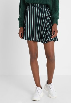 Kvinde iført en grøn sweater, sort nederdel med lyseblå lodrette striber og hvide chunky sneakers, stående mod en ensfarvet baggrund.