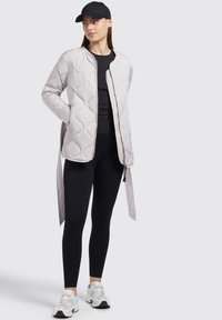 Polstret lys grå jakke med rund hals, kviltet mønster, lynlås foran og sidelæg, parret med sorte leggings og hvide sneakers.