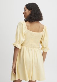 Rochie scurtă galbenă cu mâneci bufante și corset smockat, având un spate pătrat și o siluetă lejeră, realizată dintr-un material ușor.