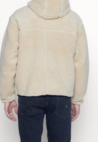 Persona che indossa un giubbotto di pile beige con cappuccio e jeans blu scuro, visto di spalle su uno sfondo bianco semplice.