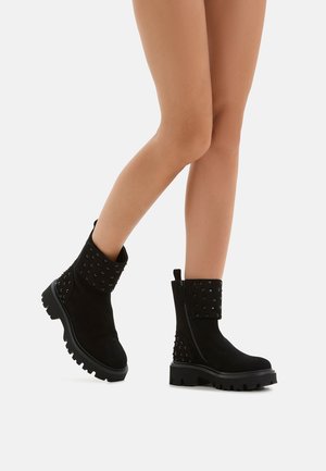 Schwarze Wildleder-Stiefeletten mit einer klobigen Sohle. Mit Nietenakzenten und einem dekorativen Bund. Glatte Textur mit einem Reißverschluss.