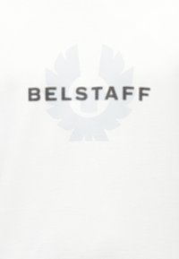 Λευκό υφασμάτινο φόντο με το κείμενο "BELSTAFF" σε μαύρη κουκκίδωση γραμματοσειρά πάνω από μεγάλο, αχνό λογότυπο με φτερωτό τροχό.