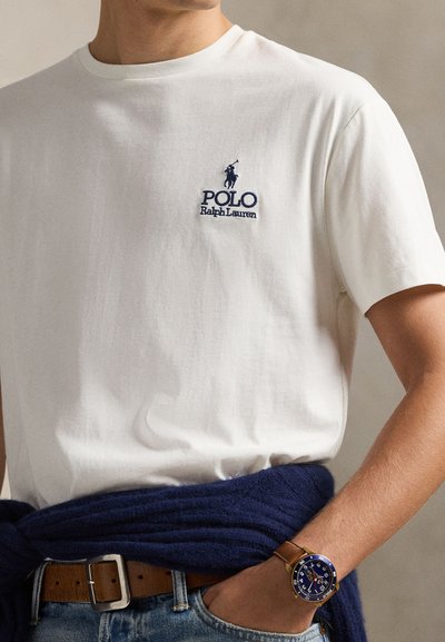 Λευκό βαμβακερό t-shirt με κεντημένο λογότυπο "Polo Ralph Lauren" σε ναυτικό μπλε, συνοδευόμενο από ναυτικό μπλε πλεκτό πουλόβερ δεμένο γύρω από τη μέση και μπλε ρολόι.