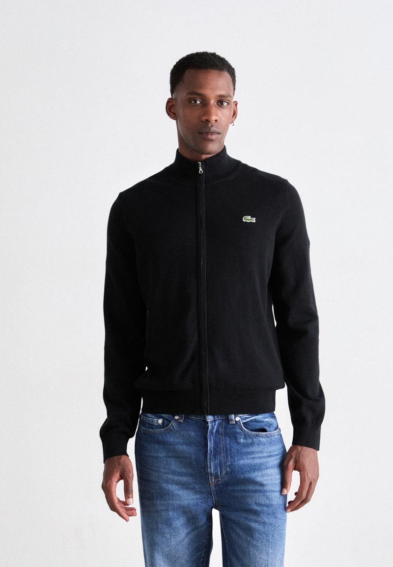 Man wearing a black zip-up sweater with a small crocodile logo and blue jeans standing against a plain white background.