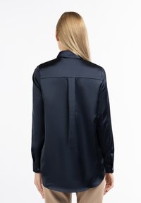Blusa di satin blu navy con maniche lunghe, colletto classico, polsini con bottoni e una piega centrale sul retro. Tessuto liscio e design leggermente allungato.