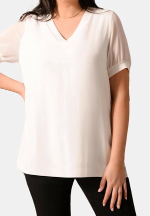 Blusa blanca de cuello en V con mangas cortas y abullonadas. Textura de tela suave, corte relajado y dobladillo curvado. Se lleva con pantalones negros.