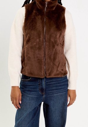 Personne portant une veste marron en fausse fourrure zippée par-dessus un pull blanc à manches longues et un jean bleu, debout avec les bras détendus.