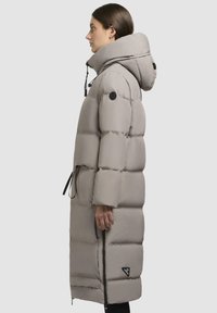 Long, light gray puffer coat with a hood, featuring a thick quilted design, side zipper, and logo patch on the sleeve.
