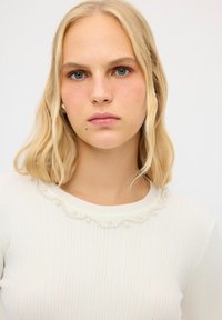 Weißer Rippstrickpullover mit einem gewellten Ausschnitt, der mit kleinen dekorativen Perlen verziert ist. Der Pullover zeigt ein enganliegendes Design und eine glatte Textur.