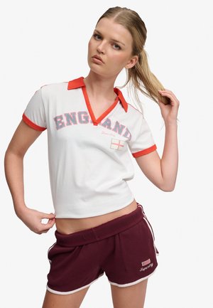 Joven mujer tirando de su coleta con una mano y ajustando su polo blanco de Inglaterra con ribete rojo y pantalones cortos burdeos.