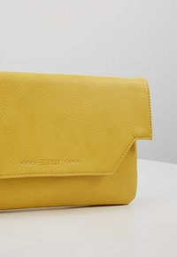 Pochette en cuir jaune avec fermeture à rabat embossée du logo Esprit, présentée sur une surface blanche sur fond neutre.