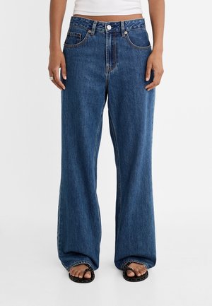 Persoon draagt losse blauwe spijkerbroek, wit cropped top en zwarte sandalen met studs, staande tegen een effen witte achtergrond.